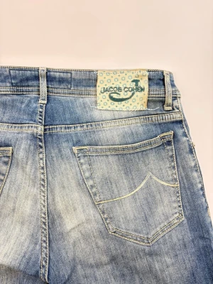 Blå jeans från Jacob Cohen - Snygga blå jeans från Jacob Cohen med klassisk femficksdesign och ljusa slitningar framtill. Jeansen har raka ben och en ljusblå patch med logga bak i midjan. Tillverkade i mjukt denimtyg som ger en avslappnad look.