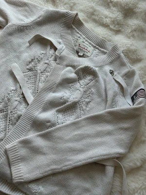 Vit kofta med brodyr från Bandelid - Snygg vit stickad kofta från Bandelid med dekorativa band och broderade detaljer framtill. Koftan har lång ärm, ribbade muddar och ett patch med amerikansk flagga på ena ärmen. Perfekt för en chill och stilren look.