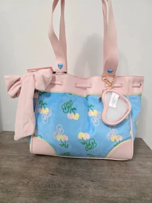 Juicy Couture daydreamer tote  - Söt handväska från Juicy Couture i pastellrosa och ljusblått med körsbärsmönster och logga. Väskan har mjukt tyg, rosa detaljer i konstläder, stor rosett och hjärtformat myntfack. Perfekt för dig som gillar gulliga accessoarer och färgglada detaljer.