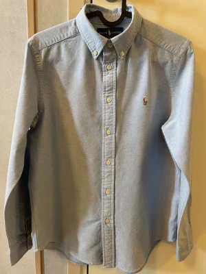Blå skjorta från Ralph Lauren - Klassisk ljusblå skjorta från Ralph Lauren med button-down krage och broderad logga på bröstet. Skjortan har långa ärmar och knäppning framtill. Perfekt för en stilren look.