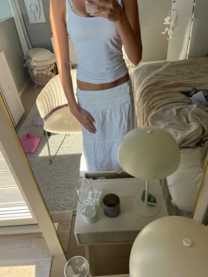 Vit långkjol med volang - Säljer en vit långkjol med bred resår i midjan och volangdetaljer. Kjolen har en luftig och flowy passform, perfekt för varma dagar. Enkel att matcha med olika toppar för en avslappnad look.