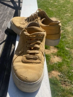 Beige sneakers med snörning - Snygga senapsgula sneakers med klassisk låg profil och tjock vit sula. Skorna har snörning framtill och är tillverkade i mocka för en trendig och avslappnad look. Perfekta för dig som gillar stilrena och unika färger.
