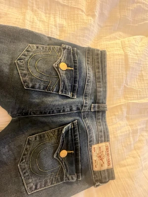 Blå True Religion jeans med kontrastsömmar - Säljer ett par blå jeans från True Religion med ikoniska kontrastsömmar och stora fickor med lock och guldfärgade knappar. Klassisk femficksmodell i tvättad denim med läderpatch bak i midjan. Dom är low Weist men håller på att få ett hål och är även uppsydda men passar om man är runt 160 cm.
