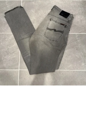 Grå jeans från Nudie Jeans - Säljer ett par grå jeans från Nudie Jeans med klassisk femficksdesign och snygg broderad bakficka. Jeansen har raka ben och är tillverkade i bomull. Perfekt för dig som gillar stilrena och tidlösa plagg.