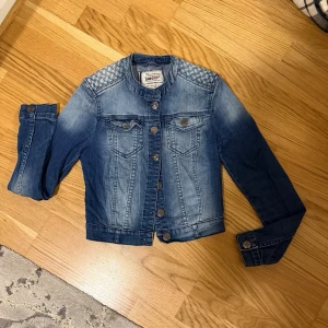 Blå jeansjacka med detaljer SHE GOVT - Snygg blå jeansjacka från SHE GOVT med coola quiltade axeldetaljer och klassiska bröstfickor med lock. Jackan har knappar framtill och vid ärmsluten samt en något kortare passform. Perfekt för dig som vill ha en trendig och tidlös look.