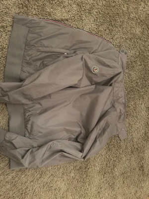 Beige vindjacka från Moncler - Snygg grå vindjacka från Moncler med klassisk logga på ärmen. Jackan har ribbade muddar, dragkedja och tryckknappar vid kragen. Tillverkad i ett lätt och smidigt material som passar perfekt för blåsiga dagar. Storlek 4, skulle säga att den passar som medium-modellen är 185 cm. Den har en liten defekt som syns på bild 4 men är inget som märks vid användning. Skick 9/10. Kom privat vid frågor!