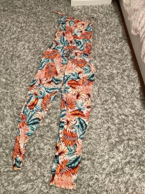 Färgglad tropisk jumpsuit med mönster - Säljer en snygg jumpsuit med tropiskt bladmönster i orange, blått och vitt. Jumpsuiten har smala axelband, markerad midja med knytband och långa ben. Perfekt för dig som gillar färg och mönster! Materialet känns lätt och luftigt.