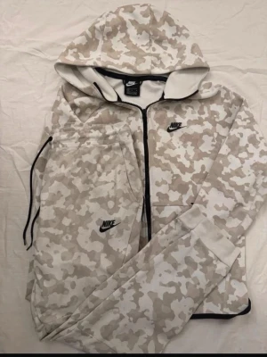 Nike mjukisbyxor beige camo, M - Nike mjukisbyxor i beige och vit camo-mönster med svart Nike-logga på benet. Byxorna har dragsko i midjan och muddar vid benslut. Tillverkade i mjukt material för maximal komfort. Perfekt för en avslappnad och sportig stil.