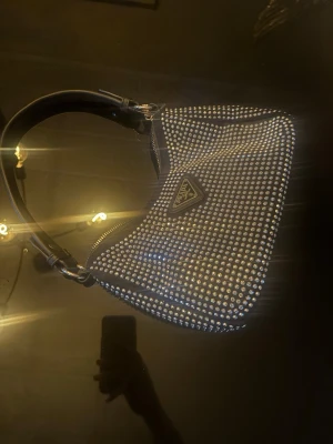 svart axelväska med strass - Snygg svart axelväska täckt med glittrande strass och klassisk trekantslogga framtill. Väskan har en böjd form och ett brett handtag i svart material. Perfekt för dig som vill sticka ut med en lyxig accessoar.