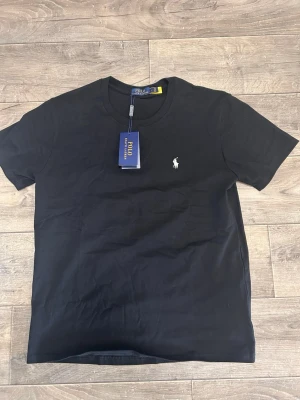 Svart t-shirt från Polo Ralph Lauren - Klassisk svart t-shirt från Polo Ralph Lauren med rund halsringning och korta ärmar. T-shirten har en liten vit broderad logga på bröstet och är tillverkad i mjuk bomull. Perfekt basplagg med stilren design.