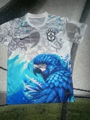 Neymar Jr Brasil tröja - Snygg fotbollströja från Nike med Neymar Jr och nummer 11 på ryggen. Framsidan har en stor blå papegoja och coola grafiska mönster i grått och vitt. Tröjan har rund hals och är kortärmad, perfekt för träning eller match. Materialet är lätt och andas.