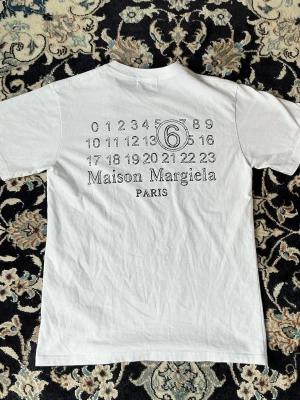 Vit Maison Margiela t-shirt med tryck - Vit t-shirt liknande Maison Margiela med ikoniskt siffertryck och logga på ryggen. Klassisk passform, rund hals och korta ärmar. Tillverkad i mjuk bomull som känns skön mot huden. Perfekt för dig som gillar minimalistisk och stilren design.