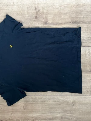 Mörkblå t-shirt med gul logga - Mörkblå t-shirt från Lyle & Scott med klassisk gul broderad logga på bröstet. T-shirten har korta ärmar och en normal passform. Perfekt basplagg till garderoben och enkel att matcha med andra kläder.
