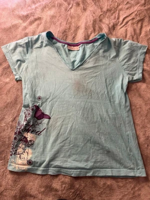 Ljusblå t-shirt med fjärilstryck - Ljusblå t-shirt med v-ringning och korta ärmar. På vänster sida finns ett stort tryck med lila fjäril, blommor och dekorativa mönster. T-shirten har en kontrastfärgad kant i halsen och är gjord i mjuk bomull. Smink fläck på framsidan, se bild. 