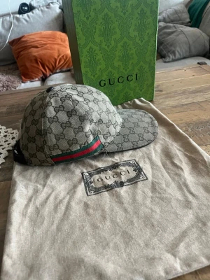 Gucci beige keps med GG-mönster - Säljer en beige keps från Gucci med det klassiska GG-monogrammet över hela kepsen. På sidan finns den ikoniska gröna och röda randen. Kepsen är i canvasmaterial och har justerbar rem bak. Perfekt för dig som vill ha en lyxig och trendig accessoar.