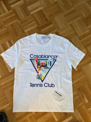 Vit Casablanca Tennis Club t-shirt - Snygg vit t-shirt från Casablanca med färgglatt tryck och texten 'Tennis Club' på bröstet. Klassisk passform och rund hals. Perfekt för dig som gillar sportig och trendig stil. Tillverkad i mjuk bomull för maximal komfort.