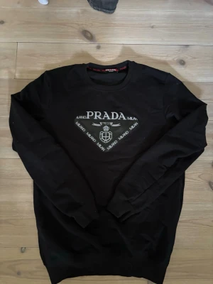 Svart sweatshirt från Prada - Svart sweatshirt från Prada med rund halsringning och tryckt logga framtill. Tröjan har långa ärmar och ribbade muddar vid ärmslut och nederkant. Perfekt för en stilren och trendig look.