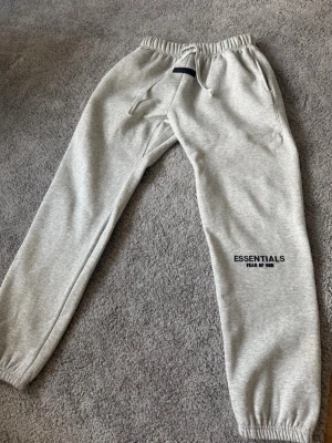 Essentials Fear of God byxor - Snygga grå mjukisbyxor från Essentials Fear of God med broderad logga på benet och patch vid midjan. Byxorna har dragsko i midjan, två sidofickor och resår vid bensluten. Tillverkade i mjukt bomullsmaterial för maximal komfort. Inprinsip aldrig använd