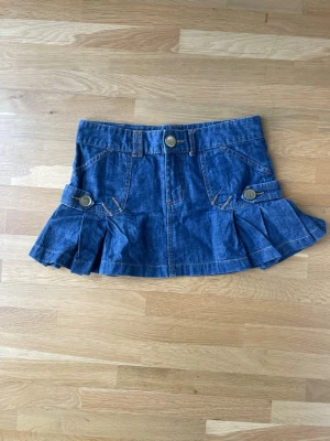 Blå jeanskjol med knappar - Snygg blå jeanskjol med dekorativa knappar och veckade detaljer på sidorna. Kjolen har fickor fram och bak samt hällor för skärp. 