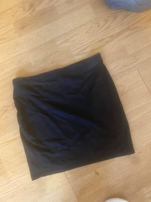 Svart enkel kortkjol - En svart, stilren och enkel kortkjol i mjukt material. Perfekt att matcha med en t-shirt eller hoodie för en avslappnad look. Kjolen har en rak passform och är lätt att styla till olika outfits.
