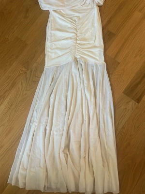 Vit långklänning med rynk och släp - Elegant vit långklänning med rynkdetalj framtill och vackert fallande kjol. Klänningen har en figurnära överdel och är tillverkad i ett glansigt material som ger en lyxig känsla. Perfekt för speciella tillfällen.