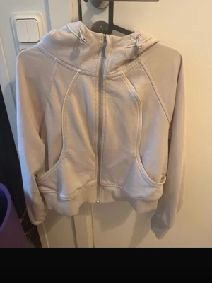 Krämfärgad zip-hoodie med sömdetaljer - En stilren krämfärgad hoodie med dragkedja framtill och markerade sömdetaljer. Hoodien har två fickor på sidorna, ribbade muddar och en klassisk huva. Perfekt för en avslappnad look och enkel att matcha med jeans eller mjukisbyxor.