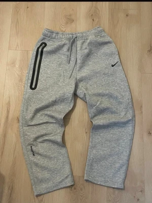 Grå mjukisbyxor från Nike x Nocta - Snygga grå mjukisbyxor från Nike x Nocta med elastisk midja och dragsko. Byxorna har en svart Nike-logga på ena benet, svart detalj vid fickan och Nocta-tryck på benet. Tillverkade i mjukt bomullsmaterial för maximal komfort.