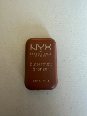 NYX buttermelt bronzer brun - NYX Professional Makeup buttermelt bronzer i en smidig, rektangulär brun plastförpackning. Bronzern har en varm brun nyans och en silkeslen formula som smälter in i huden. Perfekt för att skapa en solkysst look och definiera ansiktets konturer.