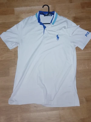 Vit pikétröja från Polo Ralph Lauren - Vit pikétröja från Polo Ralph Lauren med blå logga broderad på bröstet och blå text på ärmen. Kragen har blå och turkosa ränder. Klassisk passform och korta ärmar, perfekt för en stilren look.