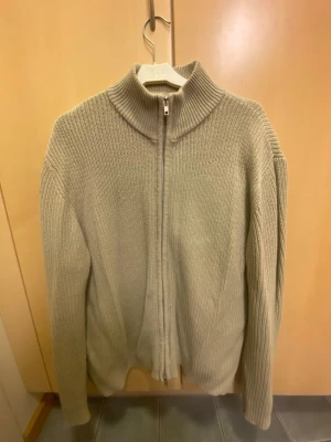 Beige stickad ziptröja - Säljer en beige stickad ziptröja med hög krage och långa ärmar. Tröjan har en grovstickad struktur och dragkedja framtill som gör den enkel att ta på och av. Perfekt för lager på lager och chill stil.
