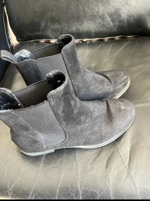 Svarta Chelsea boots i mocka - Snygga svarta Chelsea boots i mocka med klassisk rund tå och resår på sidorna. Skorna har låg klack och dragflik bak för enkel påtagning. Perfekta för dig som gillar stilrena och tidlösa skor.