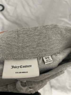Grå mjukisbyxor Juicy Couture - Grå mjukisbyxor från Juicy Couture med bred resår och dragsko i midjan. Byxorna har raka ben och två fickor framtill. Materialet är mjukt och skönt, perfekt för chill dagar. Diskret Juicy Couture-logga på ena fickan.