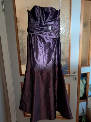 Mörklila axelbandslös klänning - Axelbandslös mörklila klänning med glansigt tyg och vacker drapering över bysten. Klänningen har en dekorativ rosett med strassdetalj i midjan och ett lätt utsvängt fall. Perfekt för dig som vill sticka ut med en elegant look.