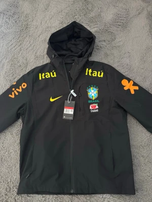 Svart Nike Brasil vindjacka med huva - Svart vindjacka från Nike med huva och dragkedja. Jackan har Brasilien CBF-logga, gula och orange sponsortryck samt Nike Swoosh på bröstet. Lätt material, meshfoder och två sidofickor. Perfekt för dig som gillar sportig stil och fotbollsinspirerade plagg.