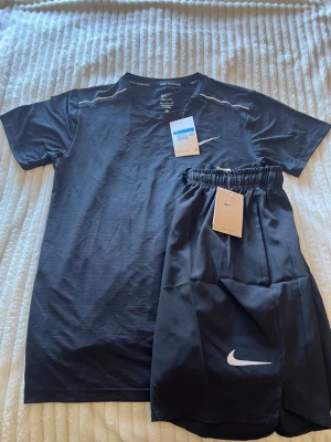 Nike Miller set - Nike Dri-FIT löparset med svart t-shirt och matchande shorts. T-shirten har diskreta ränder, rund hals och vit Nike-logga på bröstet. Shortsen är svarta med vit logga och elastisk midja. Perfekt för löpning och träning, tillverkad i funktionsmaterial.