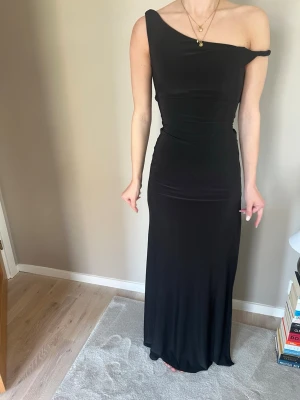 Läcker svart långklänning med slits, stl XS - Snygg svart långklänning med one shoulder-design och hög slits på sidan. Klänningen har en tight passform och är tillverkad i ett mjukt, stretchigt material som sitter skönt på kroppen. Perfekt för dig som vill sticka ut med en elegant och modern look. Använd endast vid ett tillfälle, som ny🤩! 