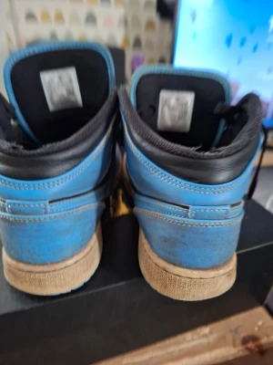 Blåa Air Jordan 1 Low sneakers - Säljer ett par blåa Air Jordan 1 Low sneakers med svarta detaljer och vit sula. Skorna har klassisk låg siluett och är tillverkade i läder med textilfoder. Perfekta för dig som gillar streetwear och vill ha en ikonisk look.