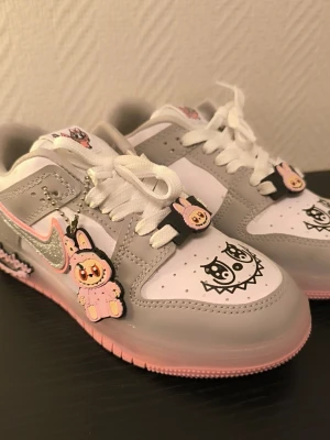 Nike sneakers med Labubu storlek 35 - Helt nya! - Unika Nike sneakers i vitt, grått och rosa med glittrig silver-Swoosh. Dekorerade med Labubu-berlocker och tecknade motiv på tån. Skorna har snörning och detaljer i syntetmaterial. Perfekta för dig som gillar lekfull och kreativ stil. Köpta i Turkiet. Aldrig använda