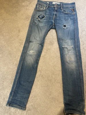 Blå slitna jeans från Replay - Säljer ett par blå anbass jeans från Replay med slitningar och hål på benen för en trendig, avslappnad look. Jeansen har klassisk femficksdesign, låg midja och är gjorda i mjukt denimtyg. Perfekta för dig som gillar streetstyle och en edgy vibe. Storlek 28/32, några tecken på användning 