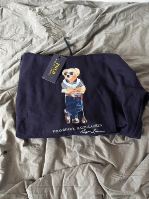 Mörkblå hoodie Polo Ralph Lauren - Snygg mörkblå hoodie från Polo Ralph Lauren med den ikoniska Polo Bear-trycket på framsidan. Hoodien har huva och är tillverkad i mjukt bomullsmaterial. Dock har den ett litet hål men ingenting märkvärdigt!
