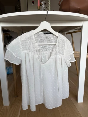 Vit blus med prickig struktur Zara - Söt vit blus från Zara med korta ärmar och v-ringning. Blusen har en lätt transparent, prickig struktur och smock upptill. Luftig och flowy passform, perfekt för sommardagar. Tunna band vid halsen för extra detalj.