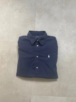 Marinblå skjorta från Polo Ralph Lauren - Klassisk marinblå skjorta från Polo Ralph Lauren med vit broderad logga på bröstet. Skjortan har knappar framtill och en stilren krage. Perfekt för dig som gillar en tidlös och clean look.