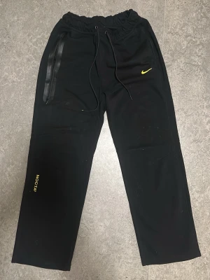Svarta NOCTA x Nike mjukisbyxor - Svarta mjukisbyxor från Nike NOCTA med gul logga och detaljer. Byxorna har resår i midjan med dragsko, raka ben och en unik ficka med dragkedja på sidan. Materialet är mjukt och bekvämt, perfekt för en avslappnad stil.