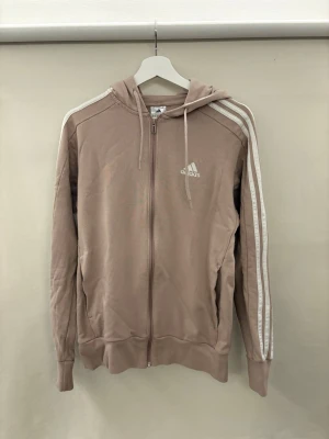 Beige hoodie med dragkedja från Adidas - Säljer en beige hoodie från Adidas med dragkedja framtill och klassiska vita ränder längs ärmarna. Hoodien har huva med dragsko och ribbade muddar. Adidas-logga i vitt på bröstet. Perfekt för en avslappnad och sportig stil. Pris kan diskuteras!
