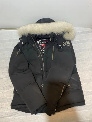 Svart pufferjacka med fuskpäls - Snygg svart pufferjacka från Moose Knuckles med avtagbar huva och fuskpäls. Jackan har flera dragkedjefickor, logga på axeln och guldig dragkedja. Perfekt för kalla vinterdagar och riktigt varm tack vare tjock vaddering.