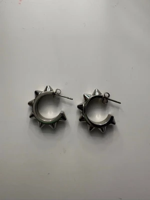 Silvriga örhängen med nitar - Coola örhängen i silverfärgad metall med rund form och utstickande nitar runt hela ringen. Perfekt för dig som gillar en edgy och unik stil. Passar till både vardagslooken och när du vill sticka ut lite extra.