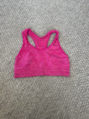 Rosa sport-bh med meshdetaljer - Säljer en snygg rosa sport-bh med racerback och meshdetaljer i ryggen. Bh:n är sömlös och har ett mönstrat tyg som ger extra ventilation. Perfekt för träning och gym. Materialet är stretchigt och bekvämt.