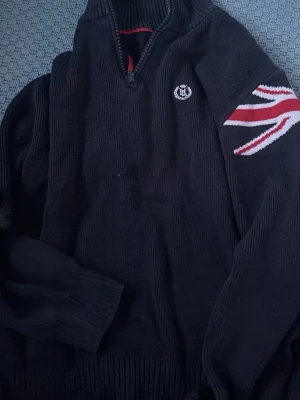 Marinblå stickad tröja Henri Lloyd - Snygg marinblå stickad tröja från Henri Lloyd med halv dragkedja, ribbade muddar och brittisk flagga på ärmen. Liten logga broderad på bröstet. Perfekt för dig som gillar klassisk och grischig  stil. 