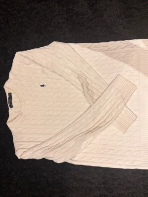 Vit kabelstickad tröja Polo Ralph Lauren - Kabelstickad vit tröja från Polo Ralph Lauren med rund halsringning och marinblå broderad logga på bröstet. Tröjan har långa ärmar och ribbade muddar. Perfekt för en klassisk och stilren look. Hör av dig vid frågor! Nypris 2099kr. Tröjan är använd ett fåtal gånger.  