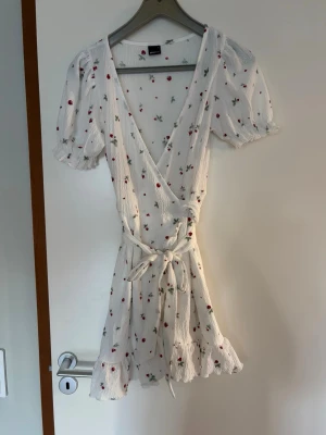 Vit omlottklänning med blommigt mönster - Supersöt vit omlottklänning från Gina Tricot med små röda blommor och gröna blad. Klänningen har kort puffärm, volangkant nertill och knytband i midjan. Perfekt för varma dagar och har en romantisk vibe. I storlek S